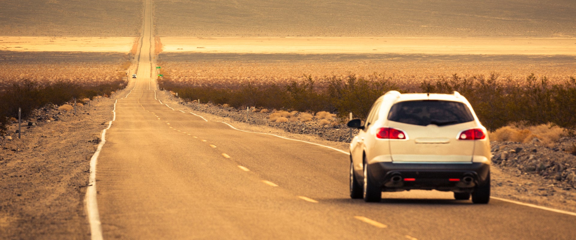 A white SUV drives down an empty, straight desert highway under a golden sunset. The scene conveys a sense of solitude and adventure.