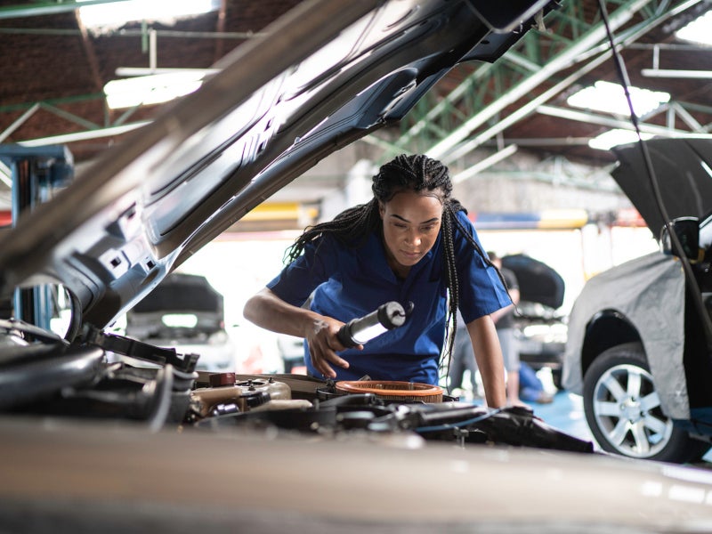 A mechanic in a blue uniform uses a flashlight to inspect a car engine in a garage, conveying focus and professionalism. Other cars are visible in the background.