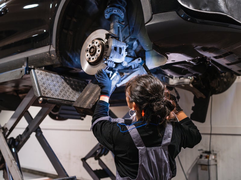 A mechanic in overalls and gloves works on a car raised on a lift, focusing on the brake system. The scene is well-lit, suggesting a professional workshop.