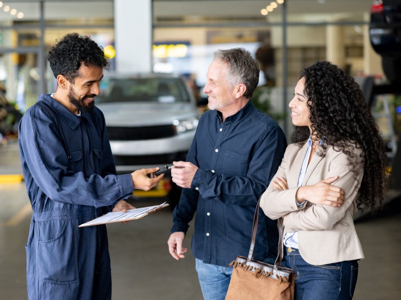 A man and woman discuss options with a mechanic at a car dealership, surrounded by vehicles and promotional materials.